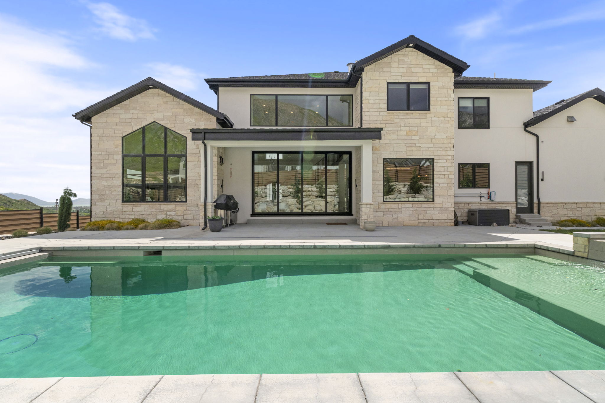 Contemporary residence featuring a rectangular swimming pool, expansive concrete patio, light-toned stone and stucco exterior, large black-framed windows, and a covered patio area