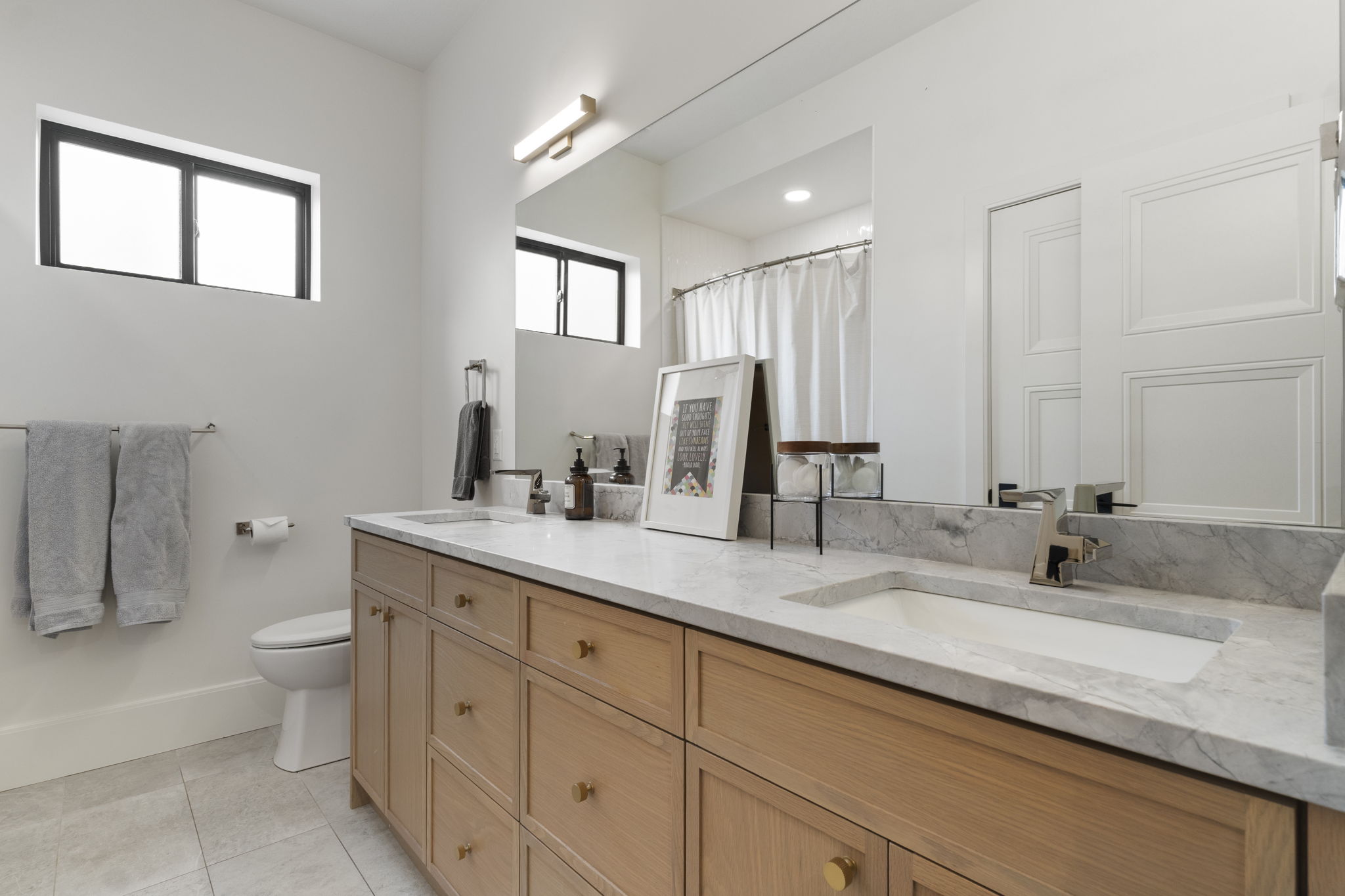 Double vanity featuring light wood cabinetry with gold-tone hardware, a light stone countertop, and two integrated rectangular sinks with modern chrome faucets