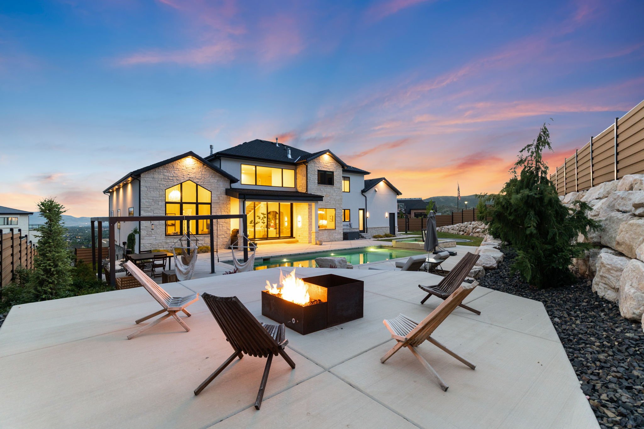 Rear exterior featuring a stone and siding facade, a swimming pool, a spacious concrete patio with a built-in fire pit, and extensive landscaping with large rock formations