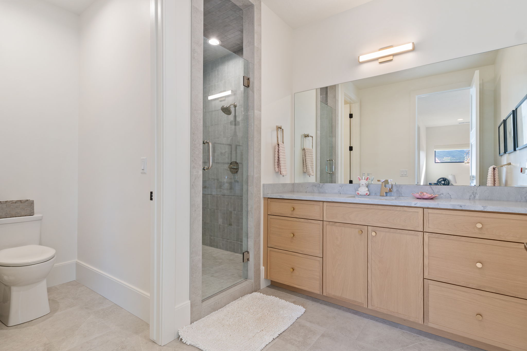 Attached contemporary bathroom featuring a light wood-finish vanity with a stone countertop, a large mirror, and an adjacent enclosed shower with tile surround