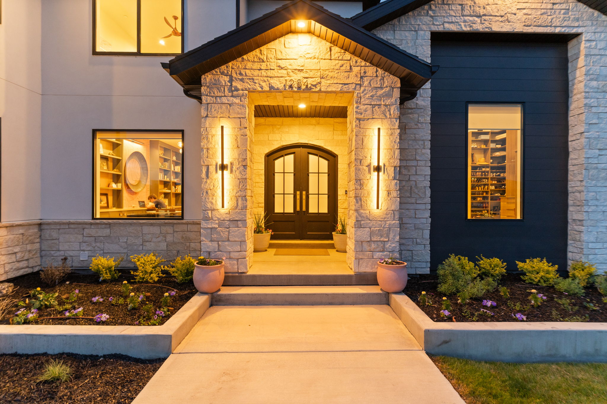 Stone and dark siding exterior with arched double entry doors, contemporary vertical sconces, a covered entryway, and concrete walkway