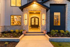 Stone and dark siding exterior with arched double entry doors, contemporary vertical sconces, a covered entryway, and concrete walkway