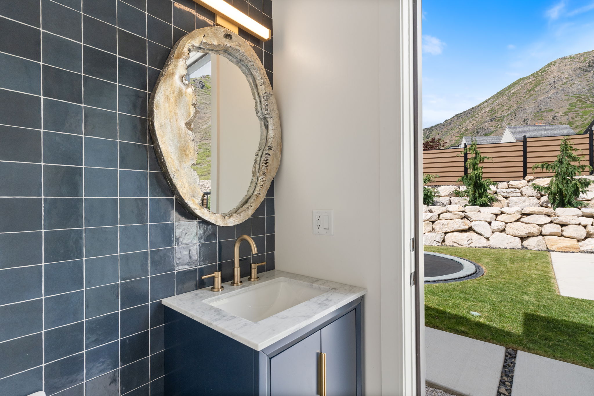 Pool bathroom vanity with a white countertop and gold-toned faucet, featuring a unique mirror and dark tiled wall