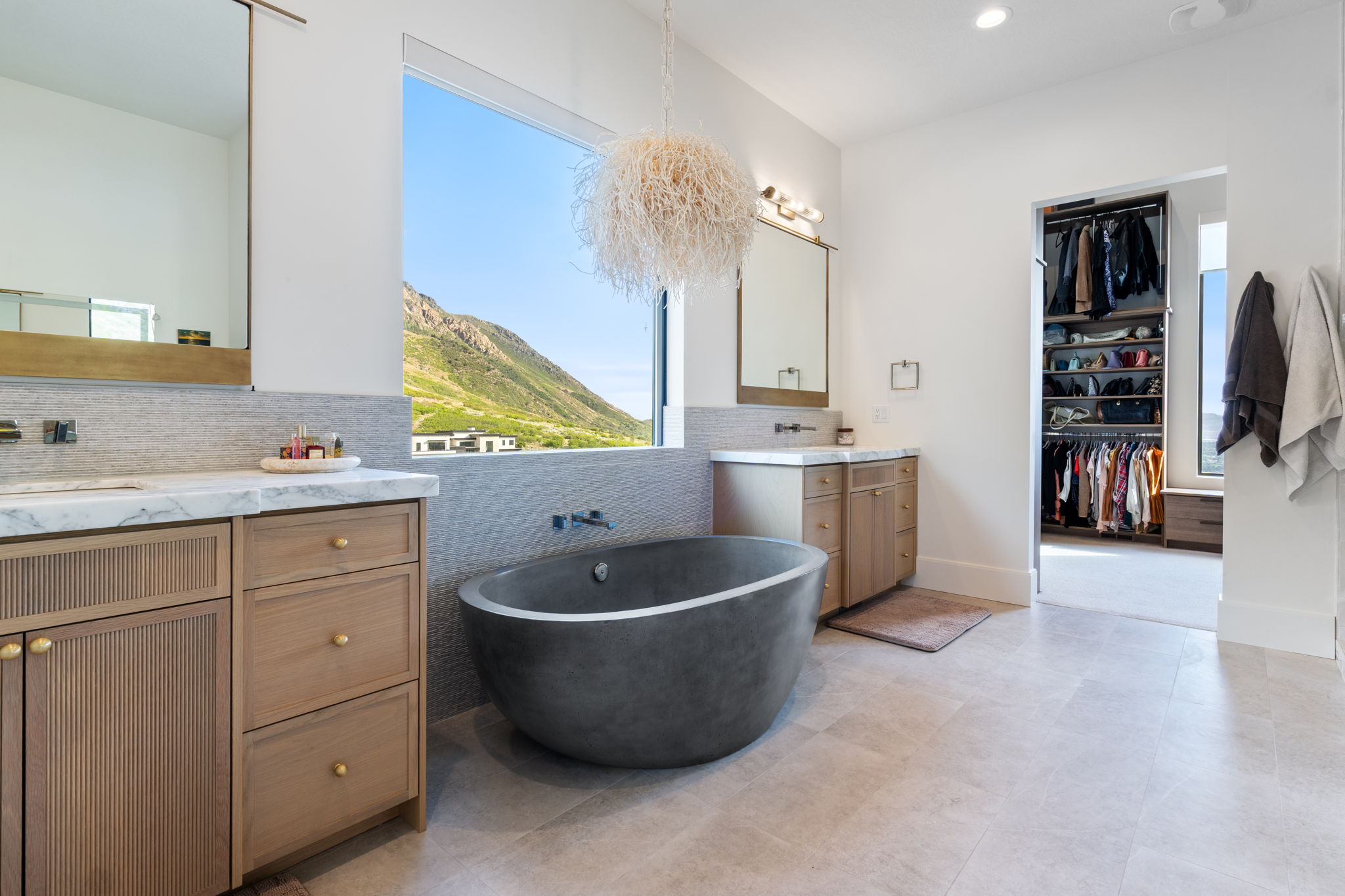 Freestanding soaking tub with a wall-mounted faucet, dual wood-finish vanities with marble countertops, large picture window with mountain scenery, and a custom built-in closet system