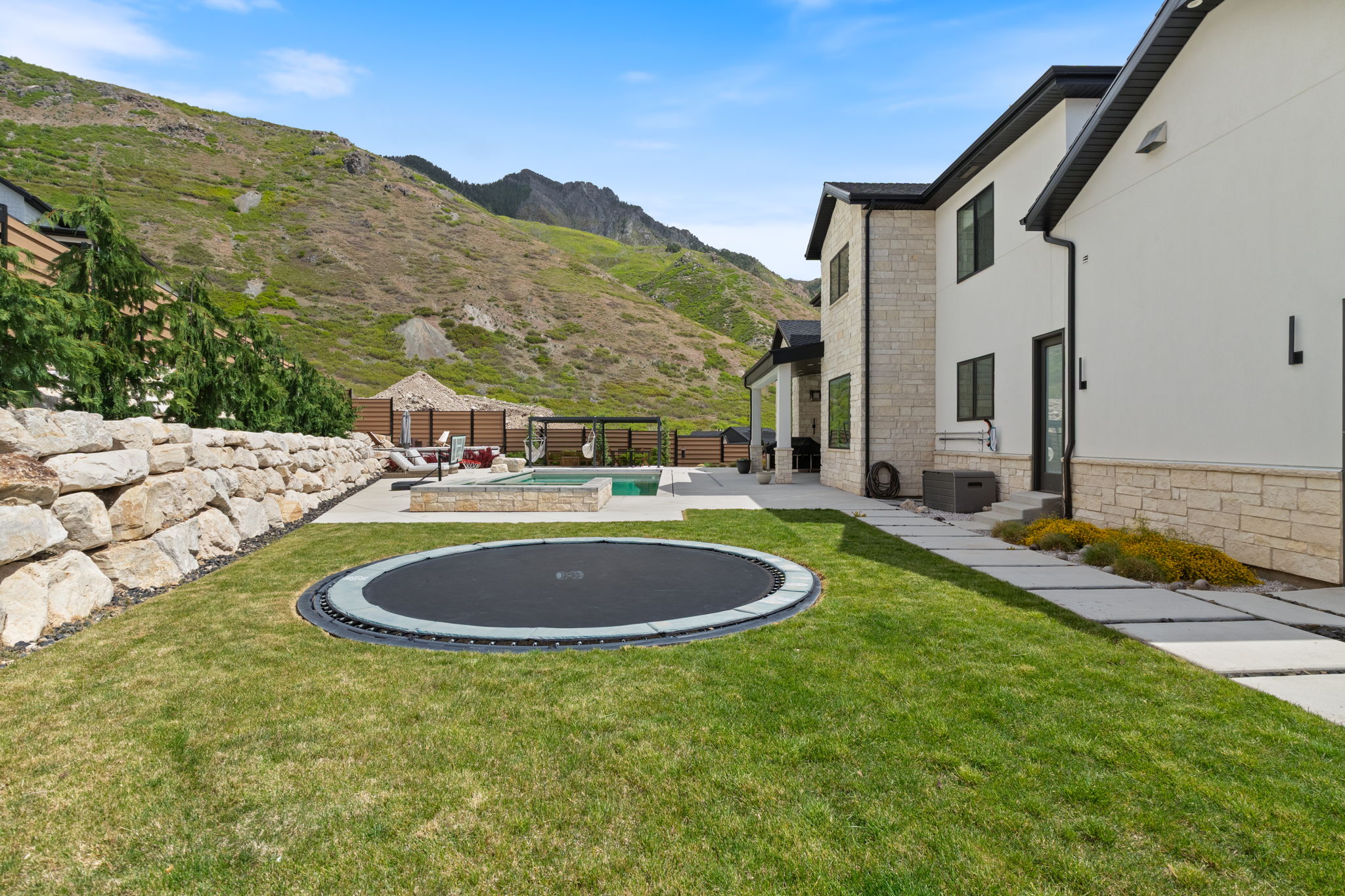 Expansive backyard featuring a manicured lawn, a built-in trampoline, concrete patio, and a retaining wall constructed of large natural stones