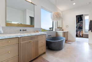 Contemporary bathroom featuring a freestanding soaking tub, dual vanity with light wood cabinetry, marble-finish countertop, and large format floor tiling