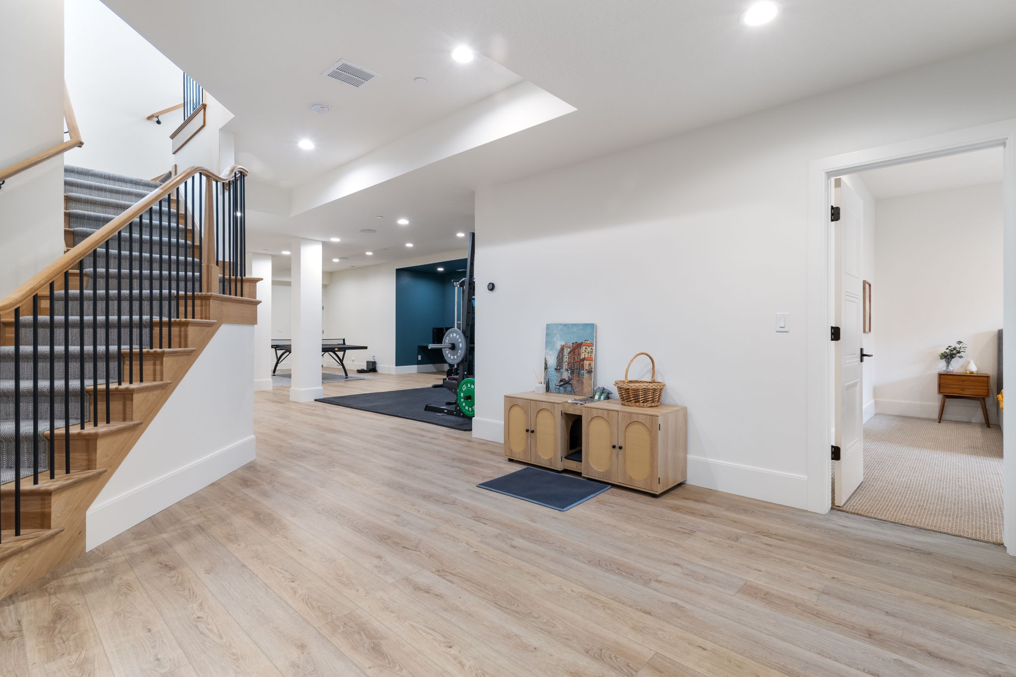 Light wood-finish flooring extending through an expansive open area featuring recessed lighting, a staircase with wood treads and black metal balusters, and a doorway opening to a carpeted space
