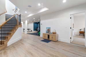 Light wood-finish flooring extending through an expansive open area featuring recessed lighting, a staircase with wood treads and black metal balusters, and a doorway opening to a carpeted space