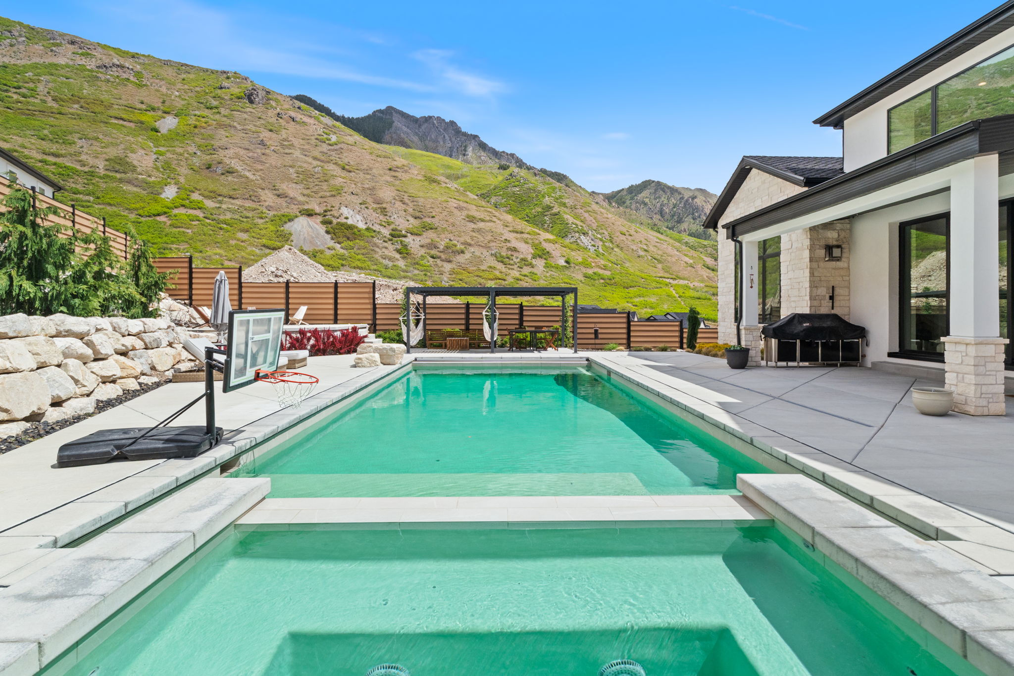 Expansive outdoor living area featuring a rectangular pool with an integrated spa, concrete patio, and stacked stone retaining wall