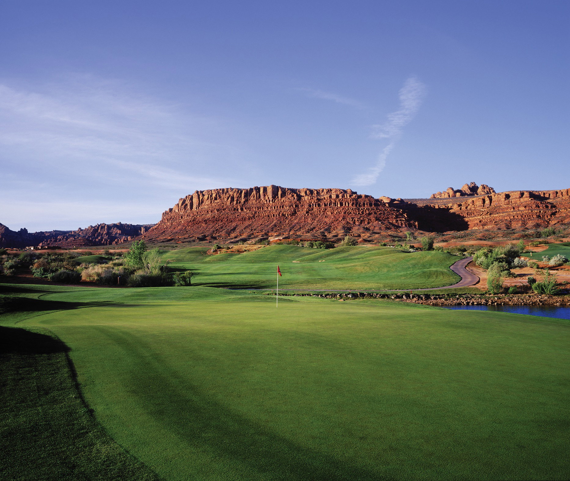 Expansive green with a red flag, bordered by a tranquil blue pond and winding cart path
