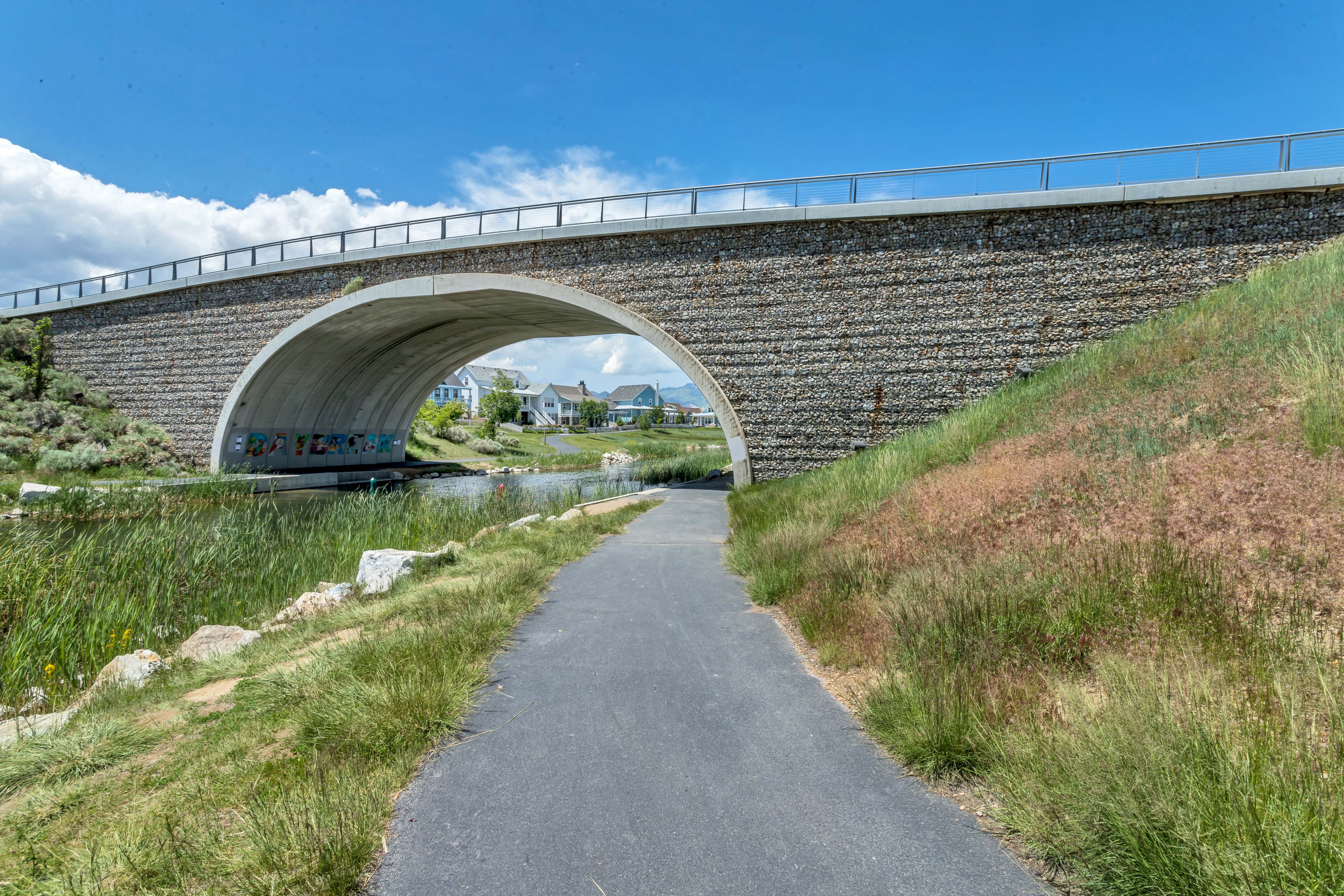 Paved pathway passing under a stone and concrete bridge, bordered by natural landscaping and a waterway