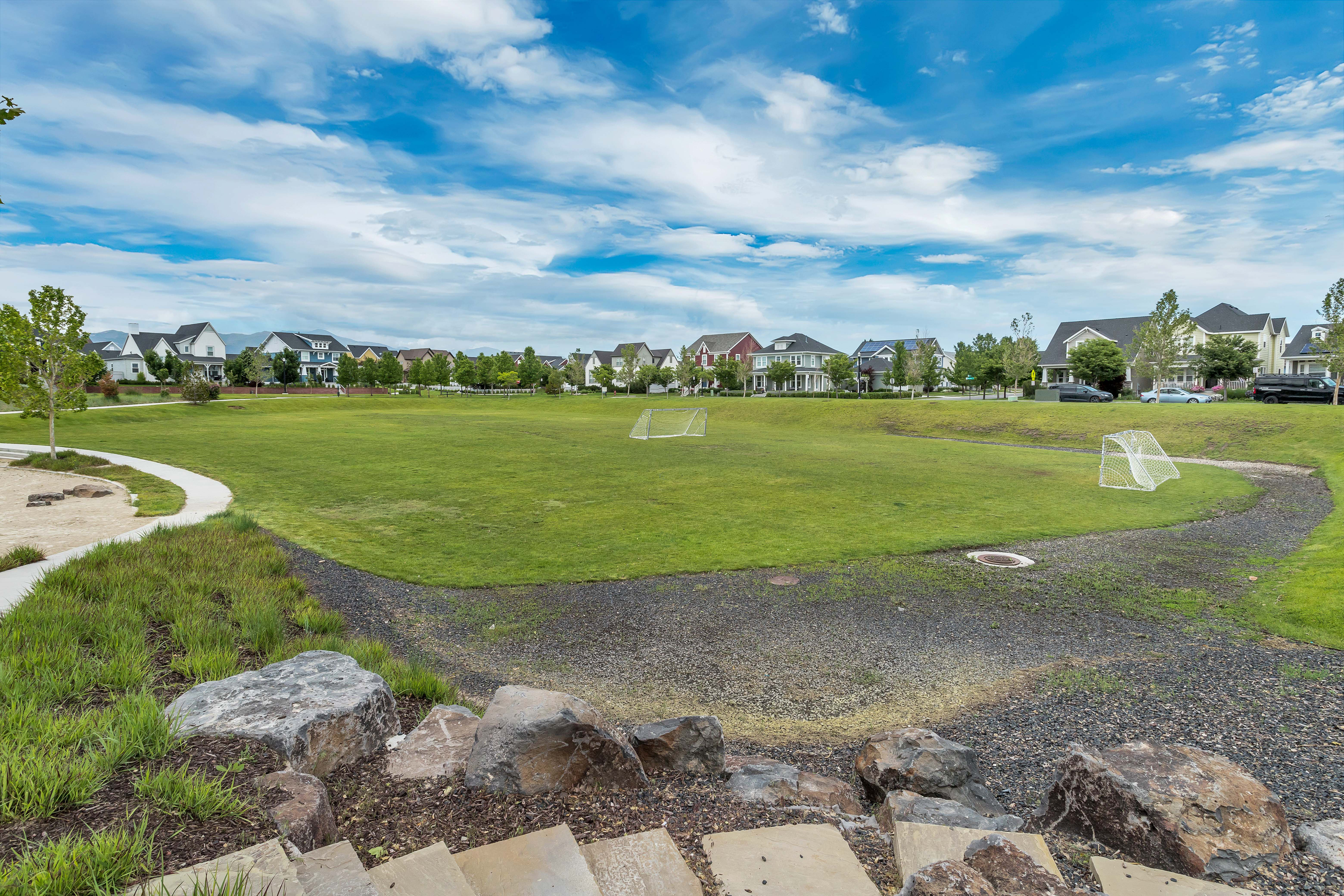 Expansive green space featuring two soccer goals, surrounded by a paved walking path and landscaped areas with mature trees