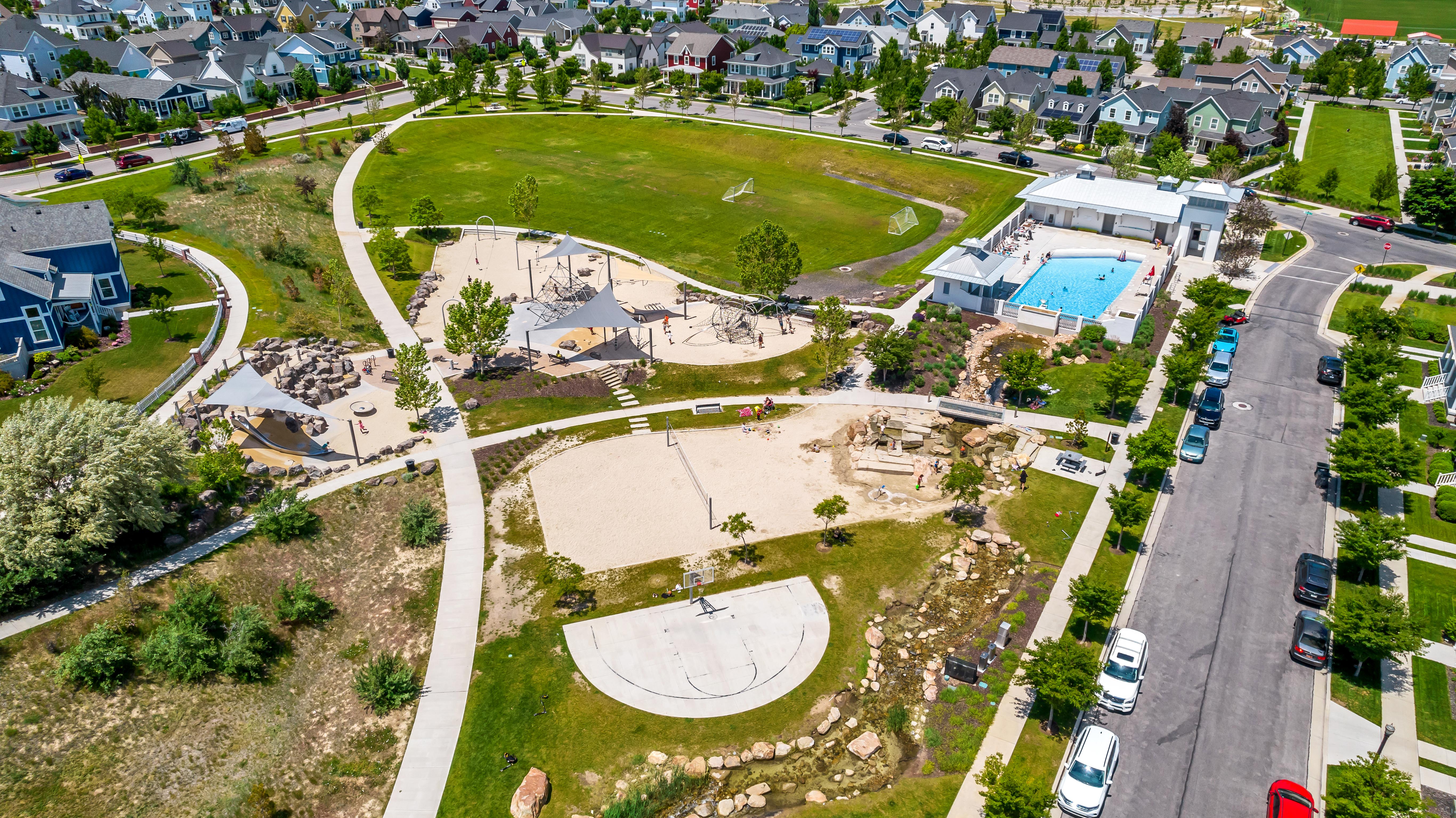Expansive community park featuring a large green space, multiple playgrounds with shade structures, a dedicated sand volleyball court, and a refreshing community pool