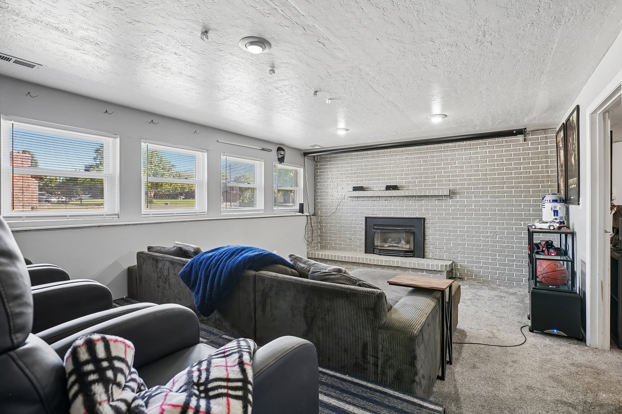 Lower-level living area featuring a light-painted brick fireplace with hearth, multiple windows, recessed lighting, and carpet flooring