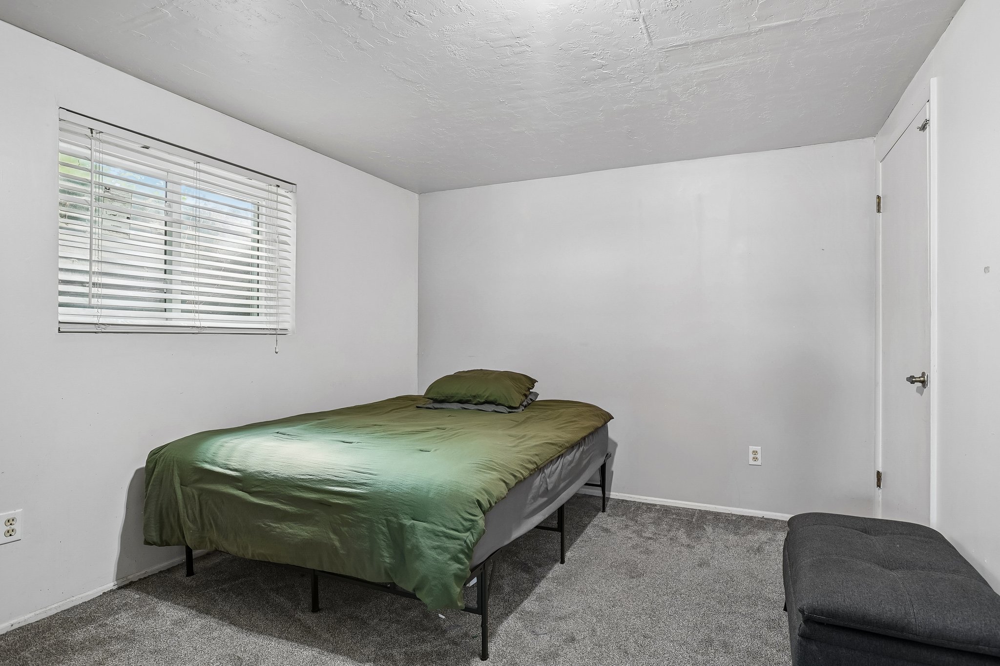 Neutral wall paint, textured ceiling, window with horizontal blinds, gray carpeting, and a white door with a black handle