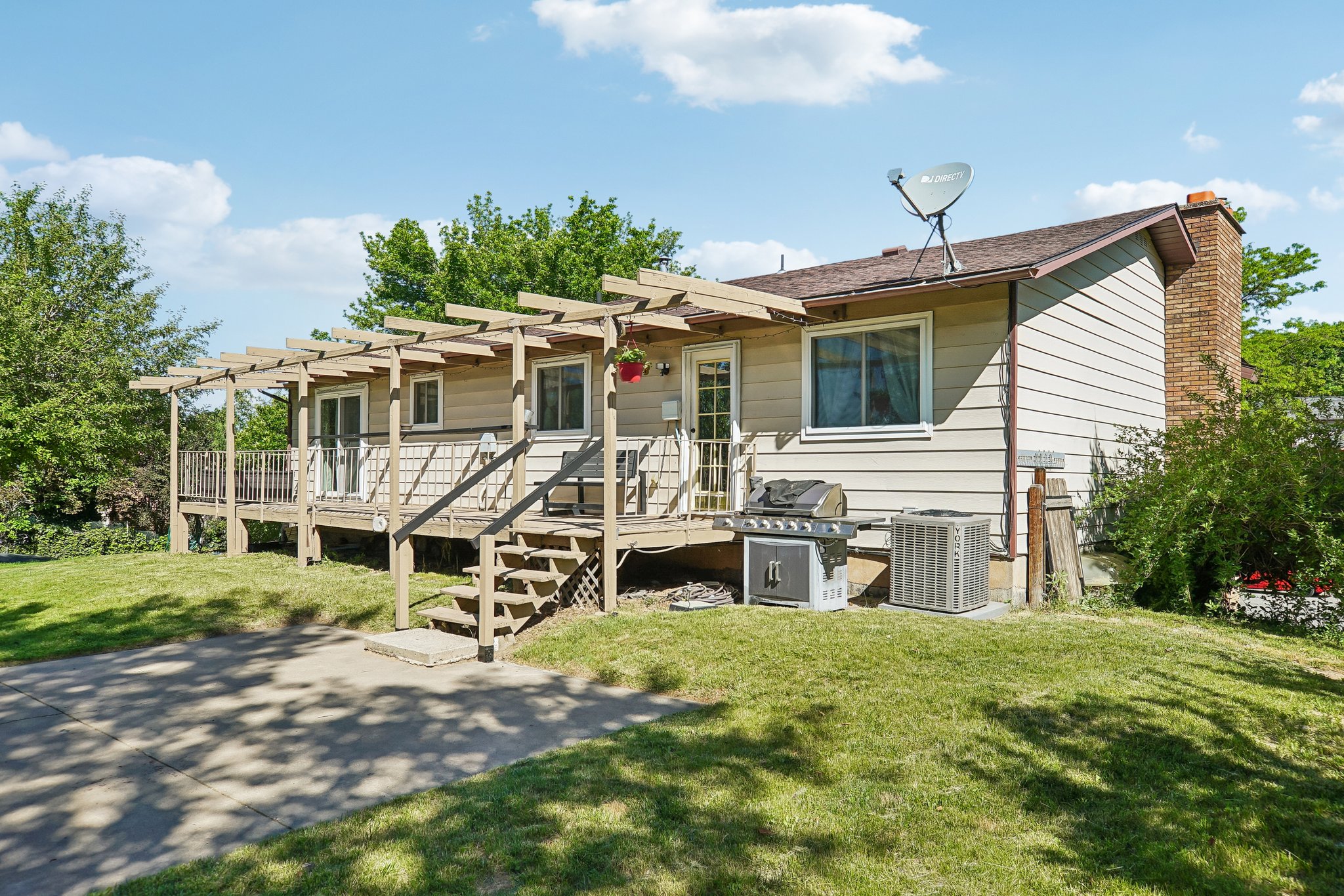 Rear exterior featuring a full-width deck with a pergola, exterior siding, a brick chimney, multiple windows, and a paved driveway