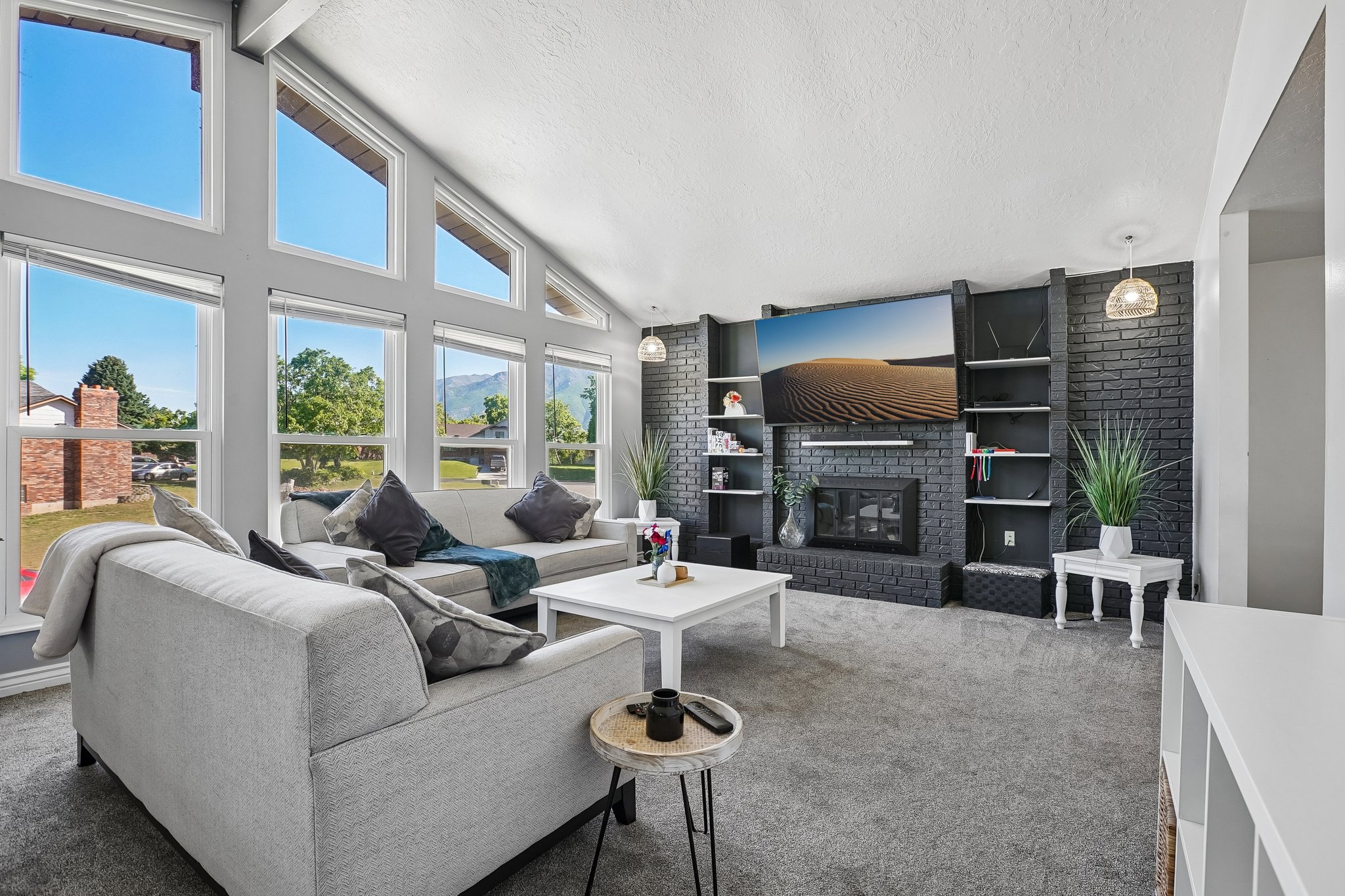 Living area featuring a vaulted ceiling with exposed beams, expansive window array, black brick fireplace surround, and built-in shelving