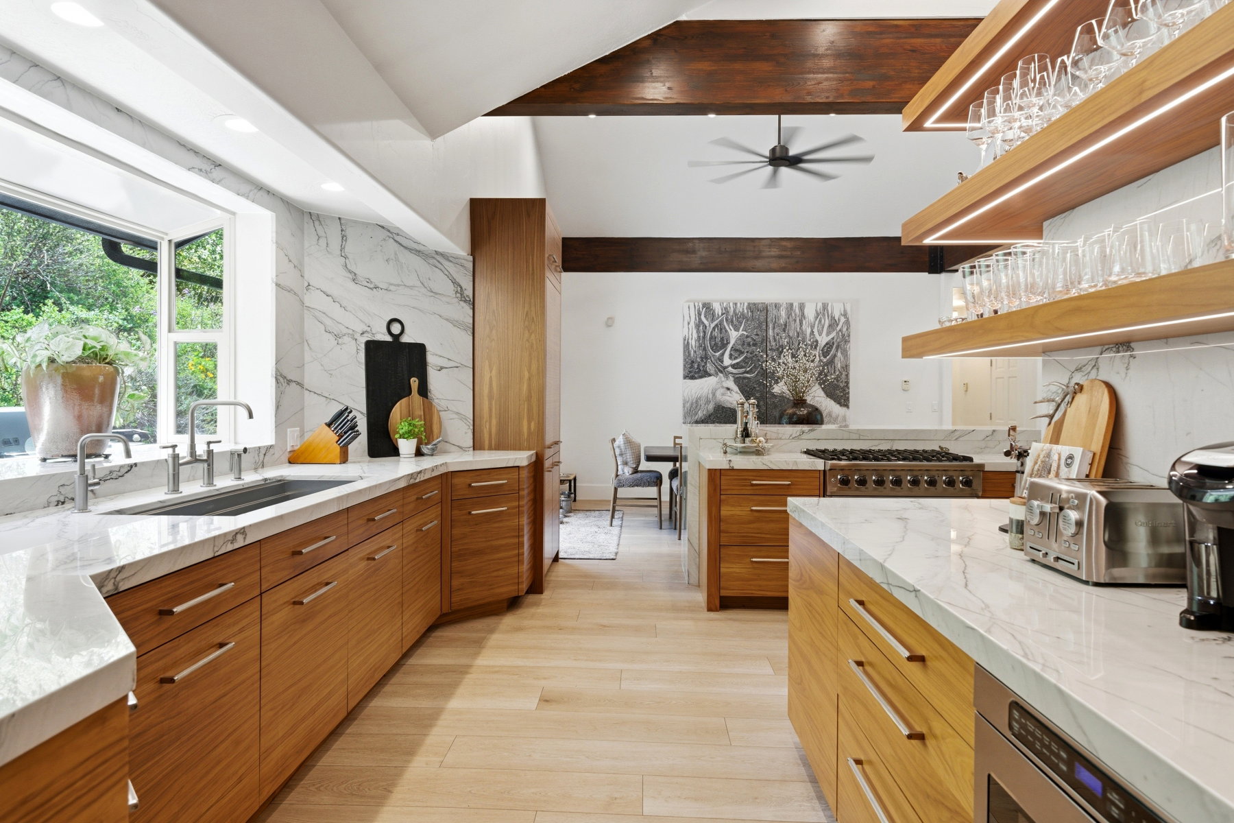 Spacious kitchen featuring a large window, light wood-finish cabinetry, marble countertops, an undermount sink, and open shelving with integrated lighting