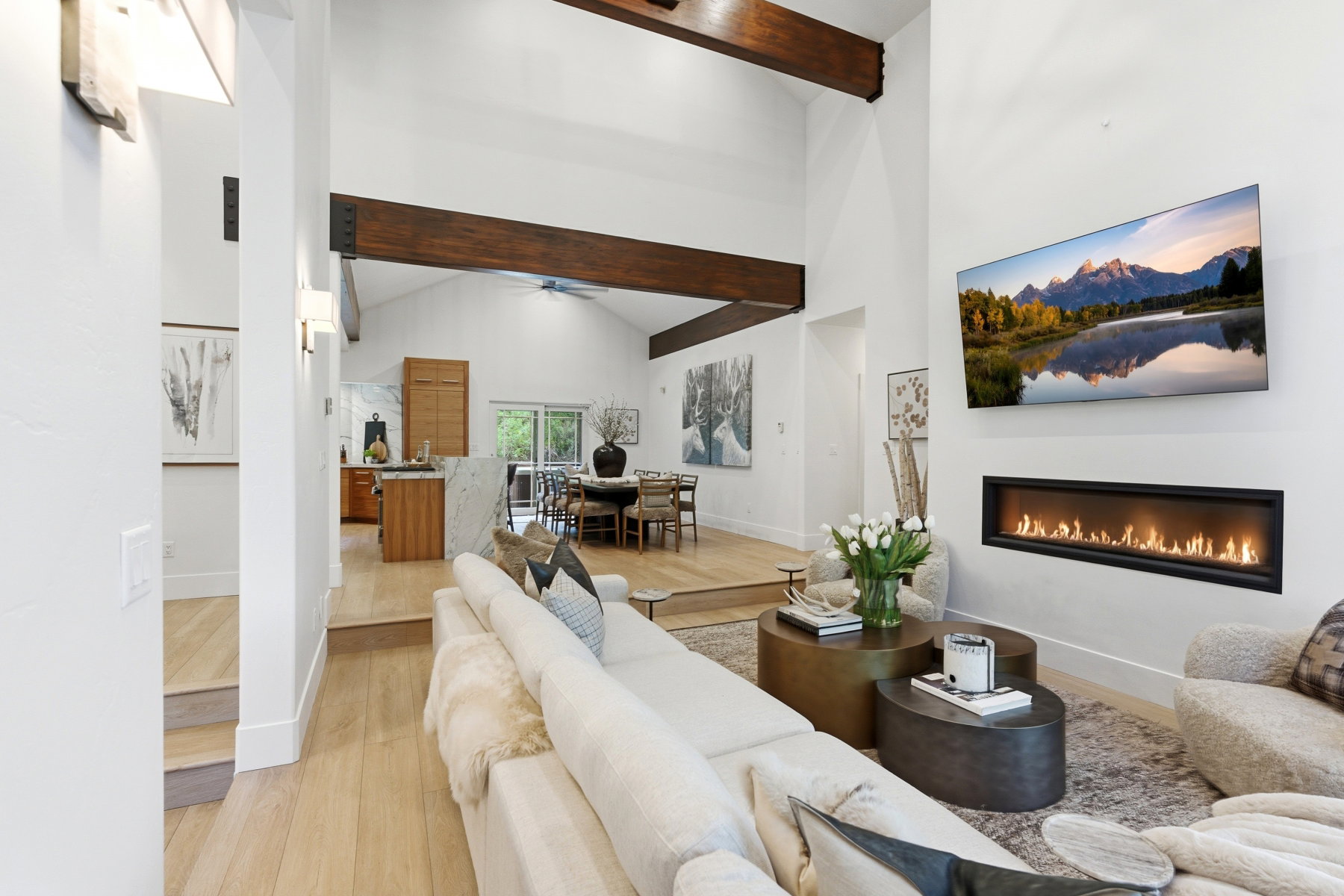 Vaulted ceilings with exposed wood beams, a linear gas fireplace, light wood-finish flooring, and a kitchen island with stone waterfall countertop
