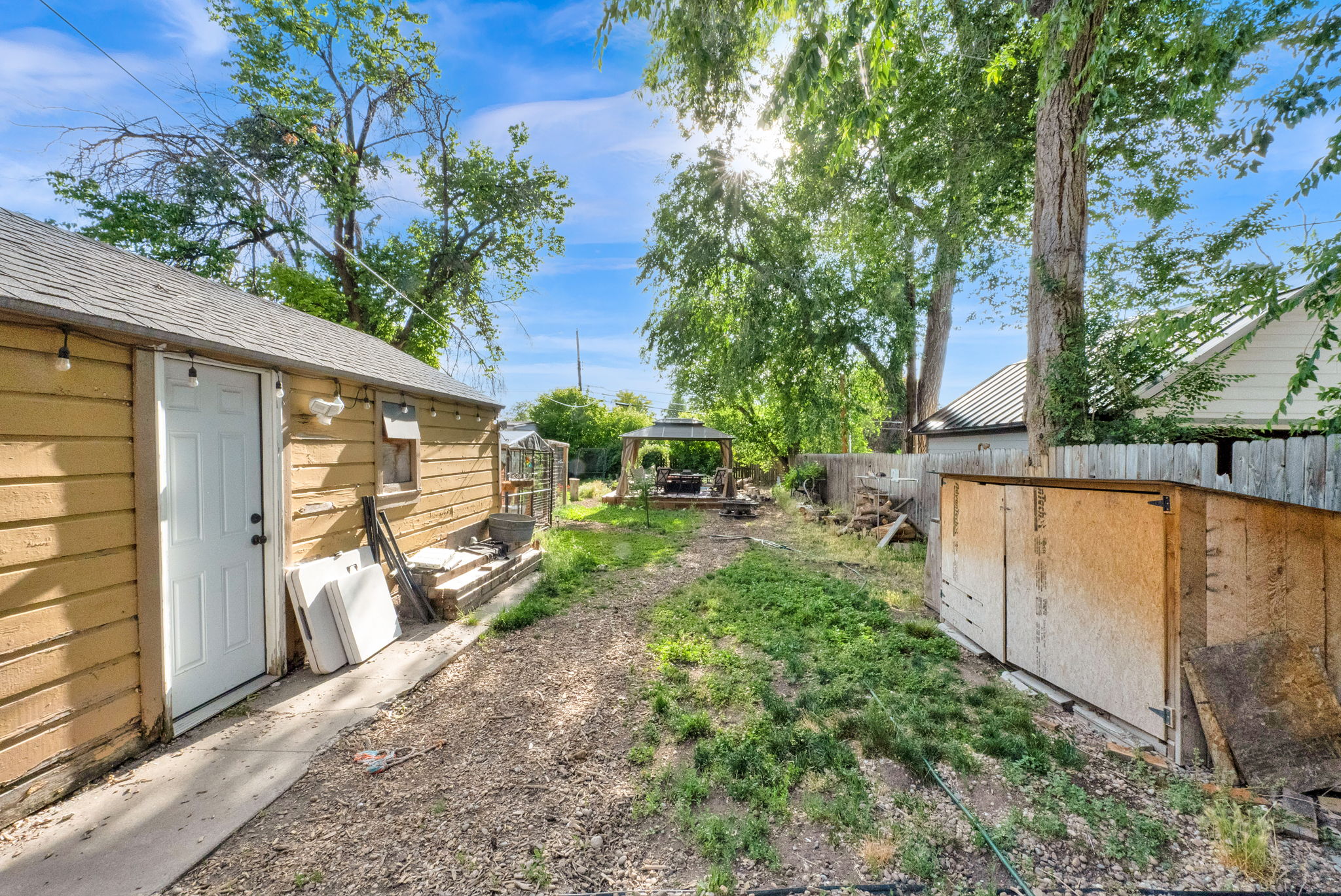 Exterior building with horizontal wood siding and white door, concrete pathway, mature trees, and a wooden fence