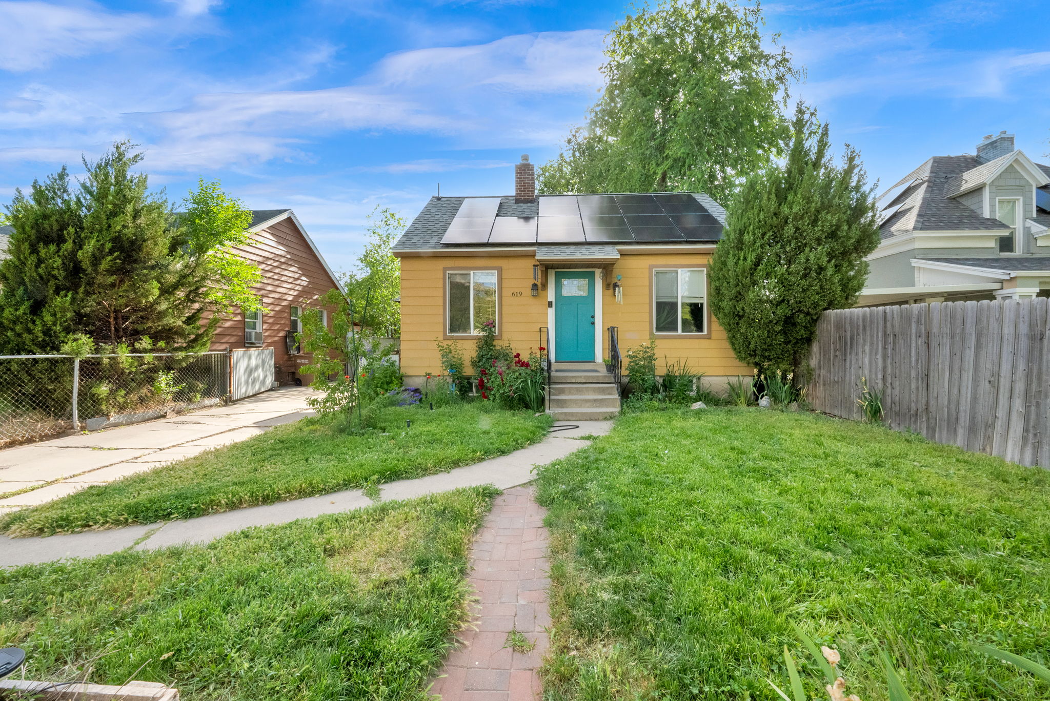 Yellow exterior with a vibrant turquoise entry door, complemented by a brick pathway and solar panels on the roof