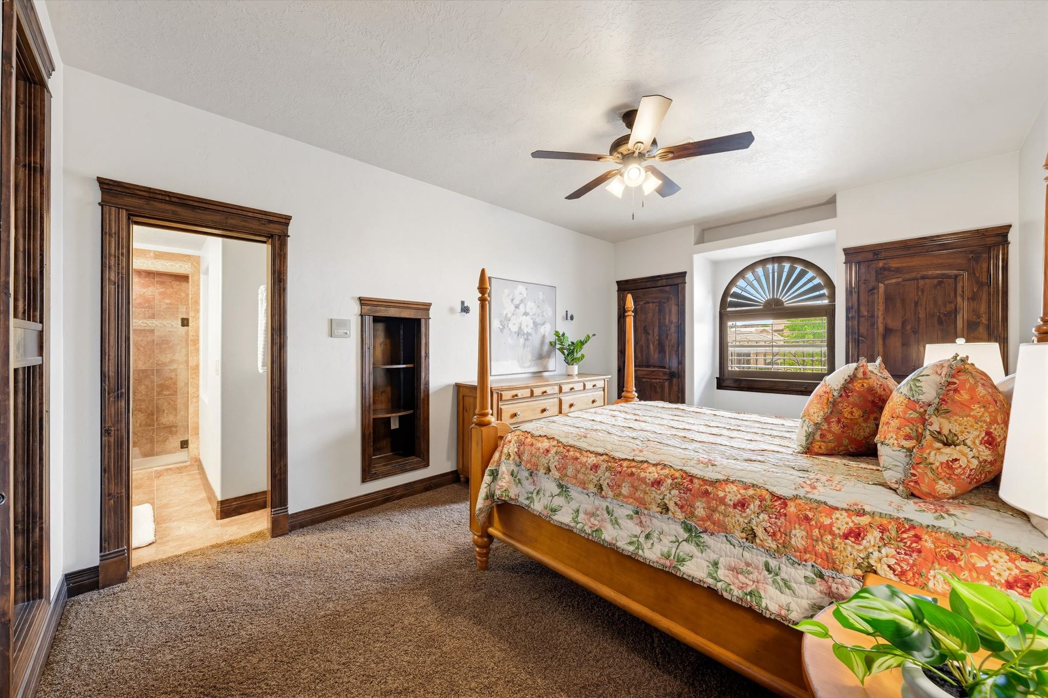 Carpeted bedroom featuring a ceiling fan, arched window with horizontal blinds, built-in recessed shelving, and a doorway opening to a bathroom with a tiled shower