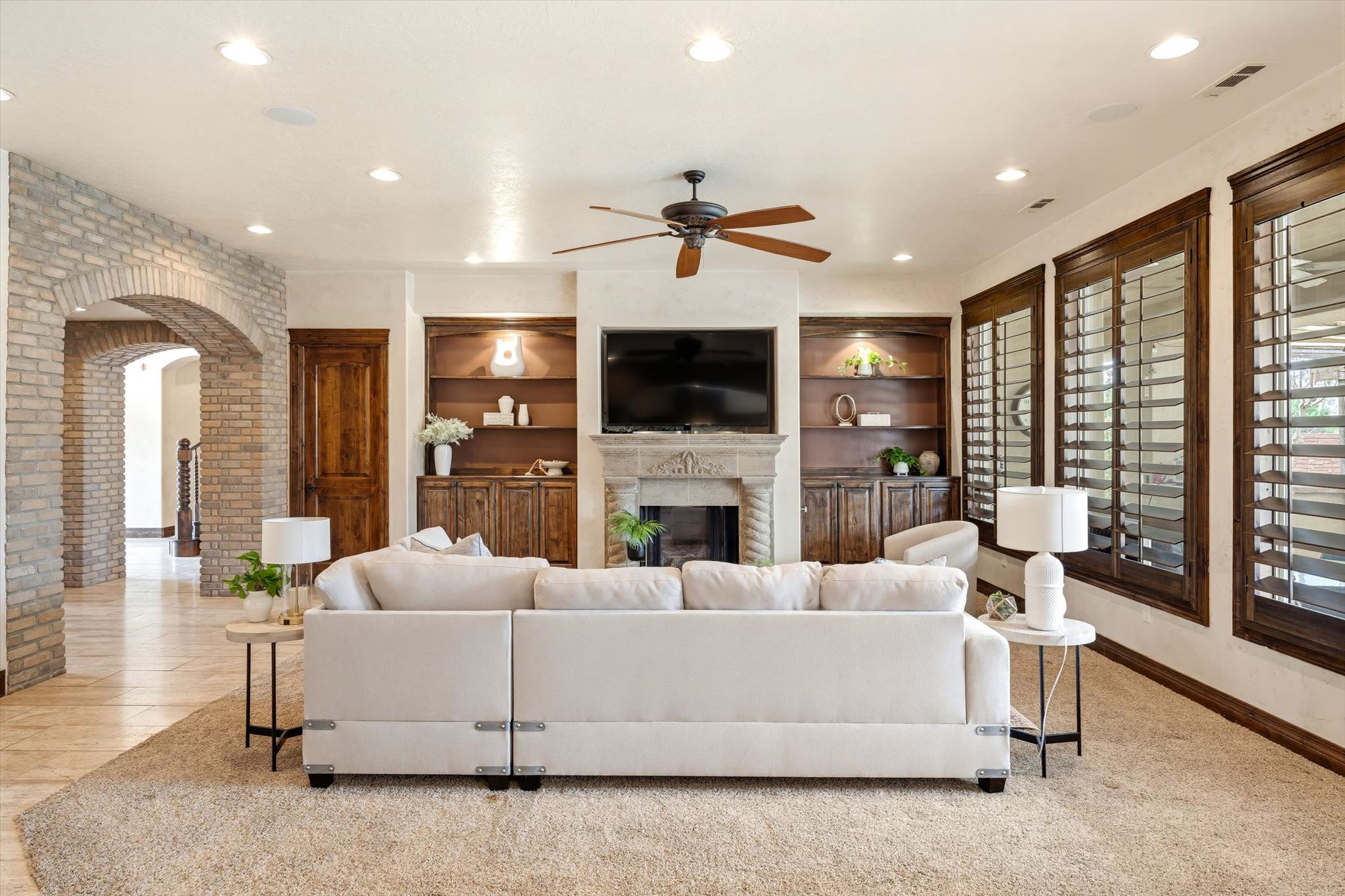 Spacious living area featuring a stone fireplace, built-in shelving, wood-finish plantation shutters, recessed lighting, and a ceiling fan