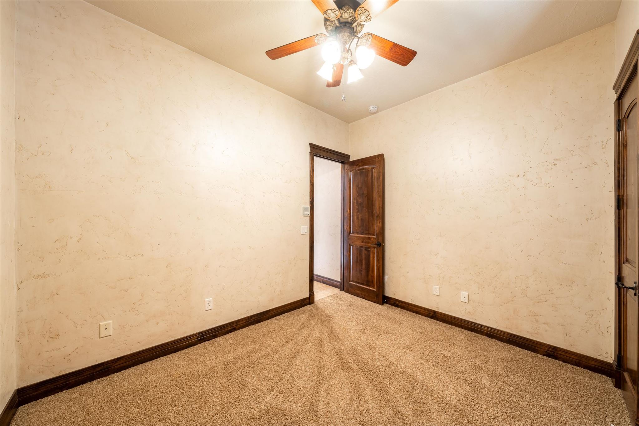 Carpeted room featuring textured walls, dark wood trim, a dark wood door, and a ceiling fan with light fixture
