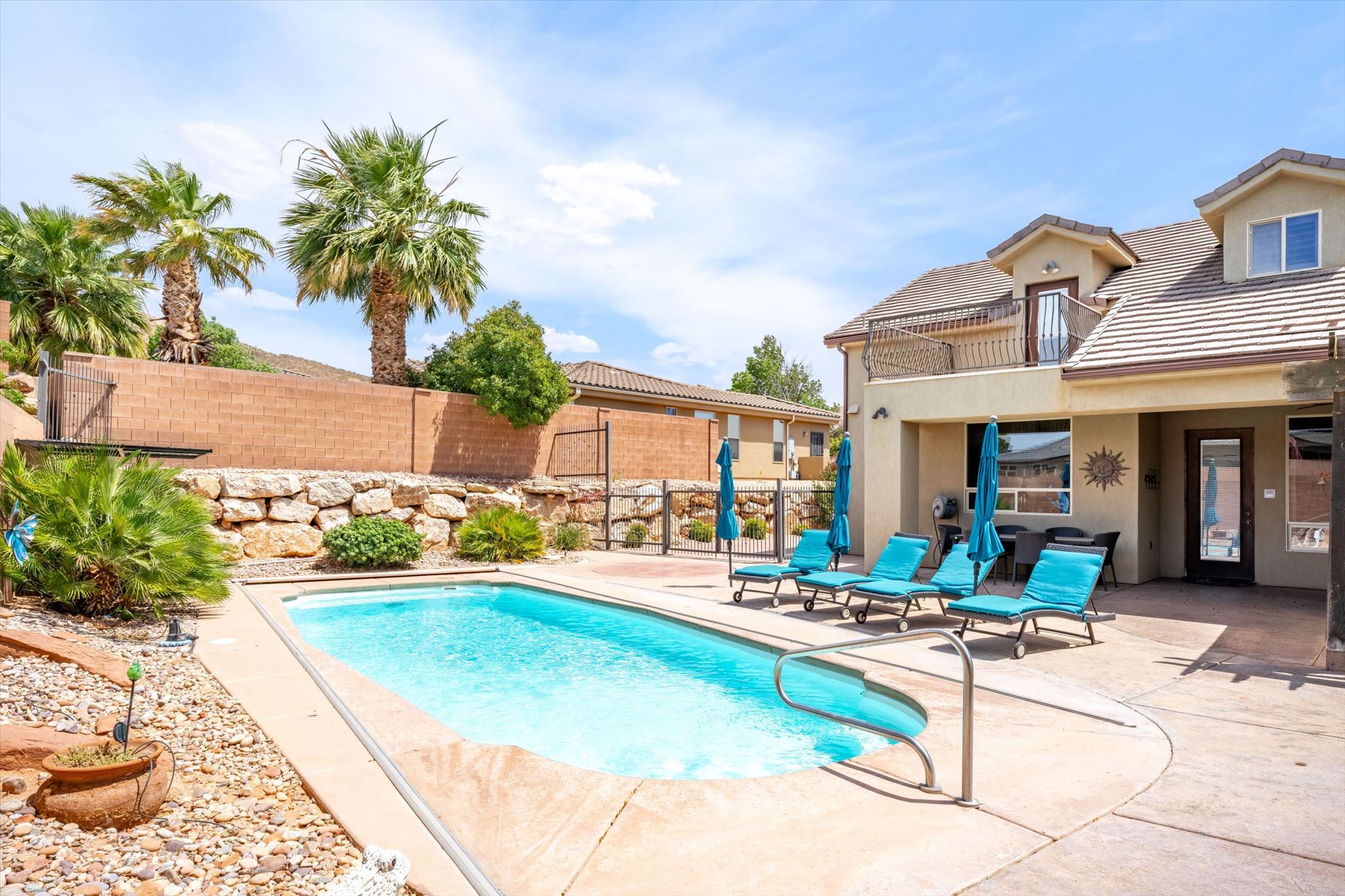 Outdoor swimming pool with concrete decking, surrounding rock landscaping, a stone retaining wall, mature palm trees, and a second-story balcony with metal railings