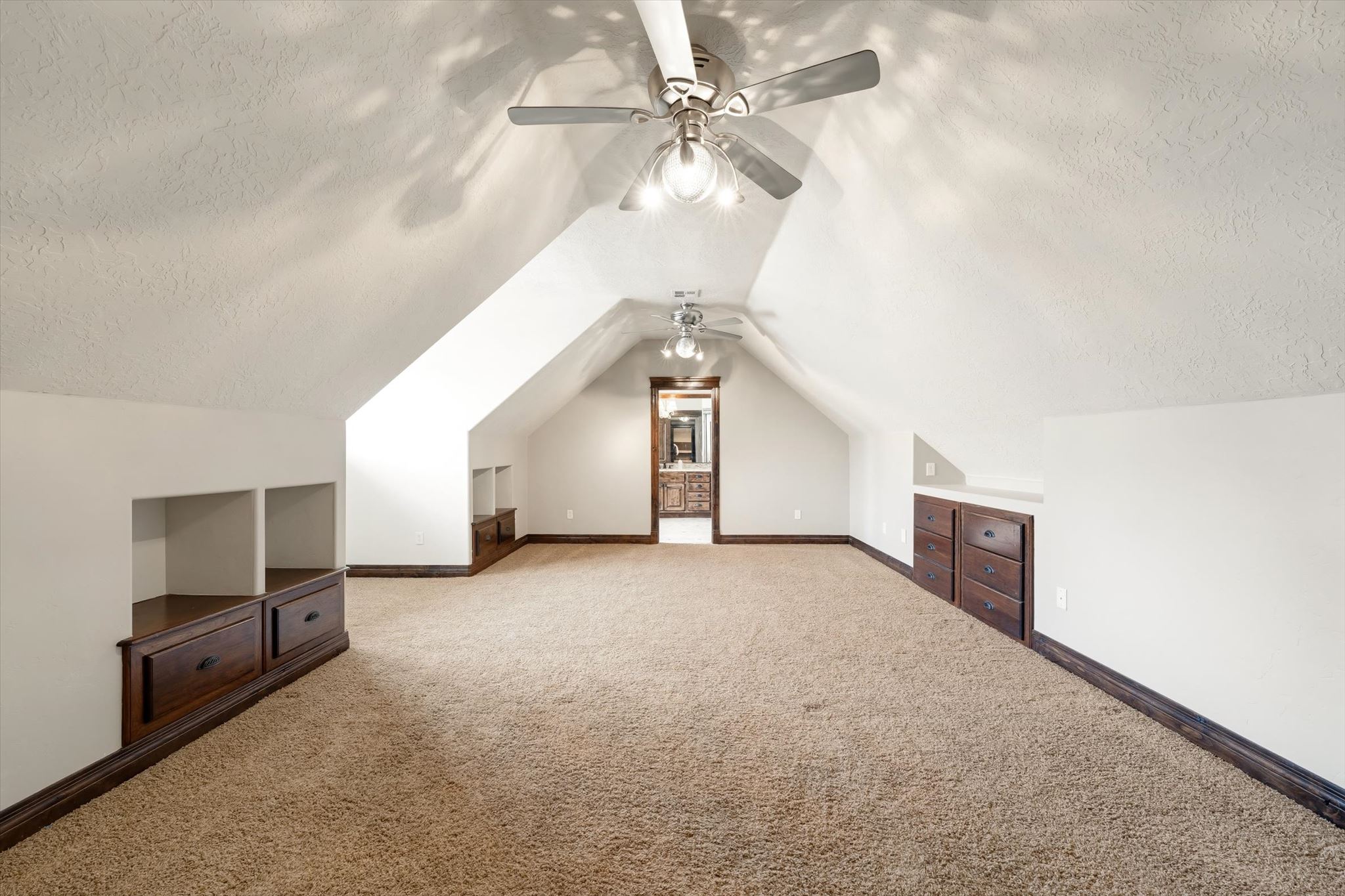 Spacious attic room featuring sloped ceilings, recessed built-in shelving, integrated wood-finish cabinetry, and two ceiling fans with integrated lighting