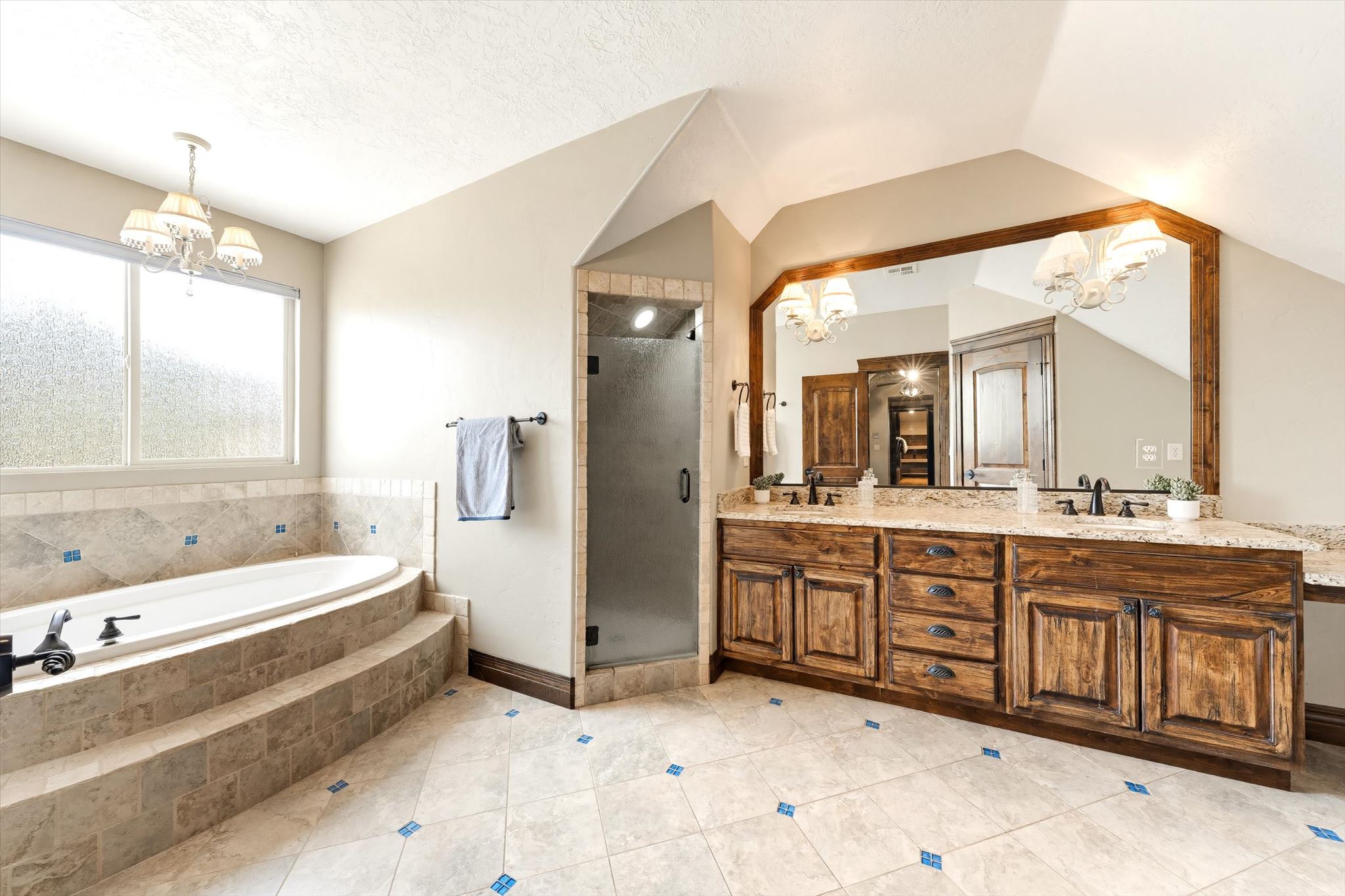 Tiled bathroom featuring a built-in soaking tub with tiered steps, an adjacent glass-enclosed shower, and a dual-sink vanity with wood-finish cabinetry and a large framed mirror