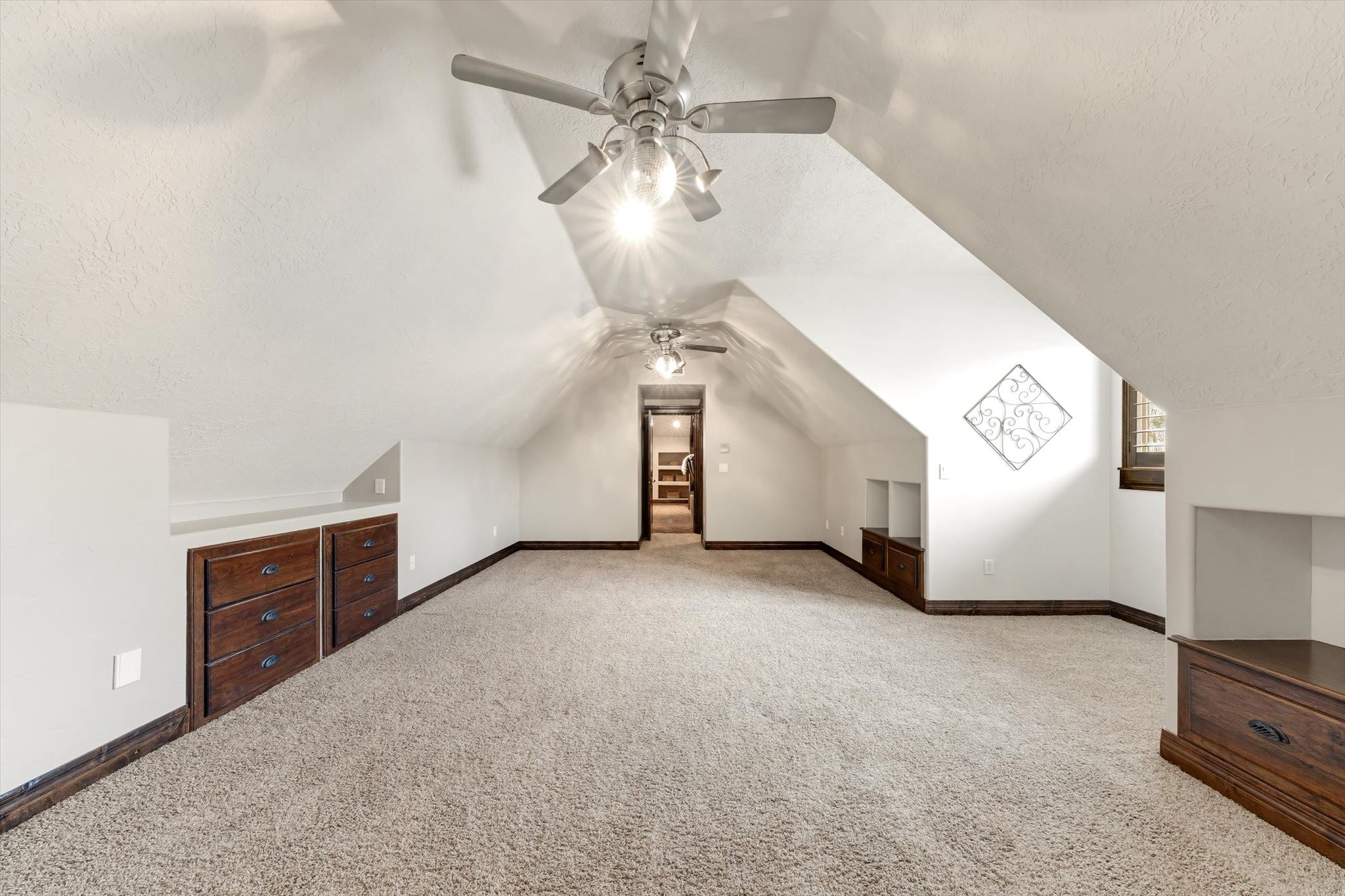 Expansive carpeted room featuring a vaulted ceiling with two ceiling fans, built-in dark wood cabinetry, recessed wall shelving, and a window with wood trim
