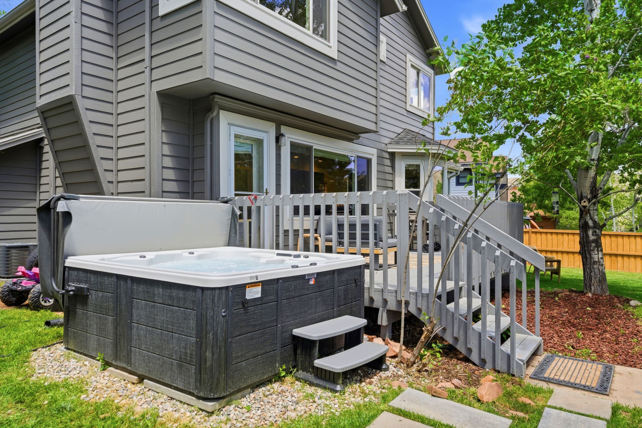 Backyard featuring a grey-sided exterior, a hot tub with steps, a wooden deck with railing and stairs, a sliding glass door, and green landscaping with mature trees