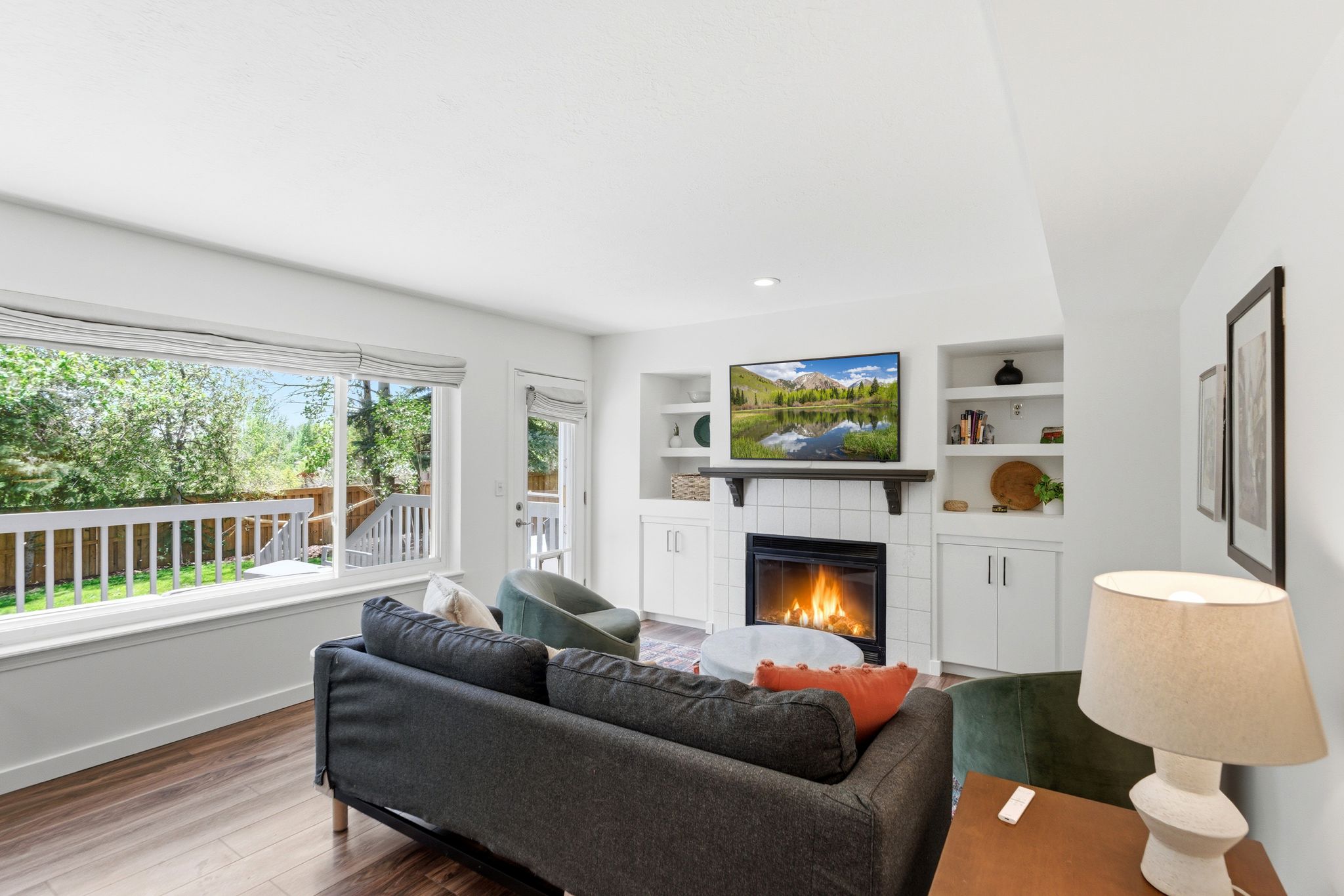 Family area featuring LVP flooring, a white tile-surround fireplace with a wood mantel, built-in shelving, and a large window overlooking a wooden deck and green backyard