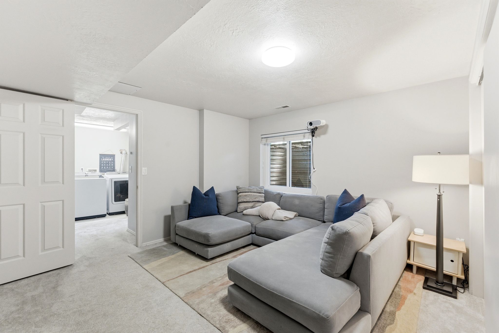 Lower Level TV/Media Room.Carpeted interior space featuring a window, built-in light fixture, and a sliding panel door opening to an adjacent laundry area with a washer and dryer