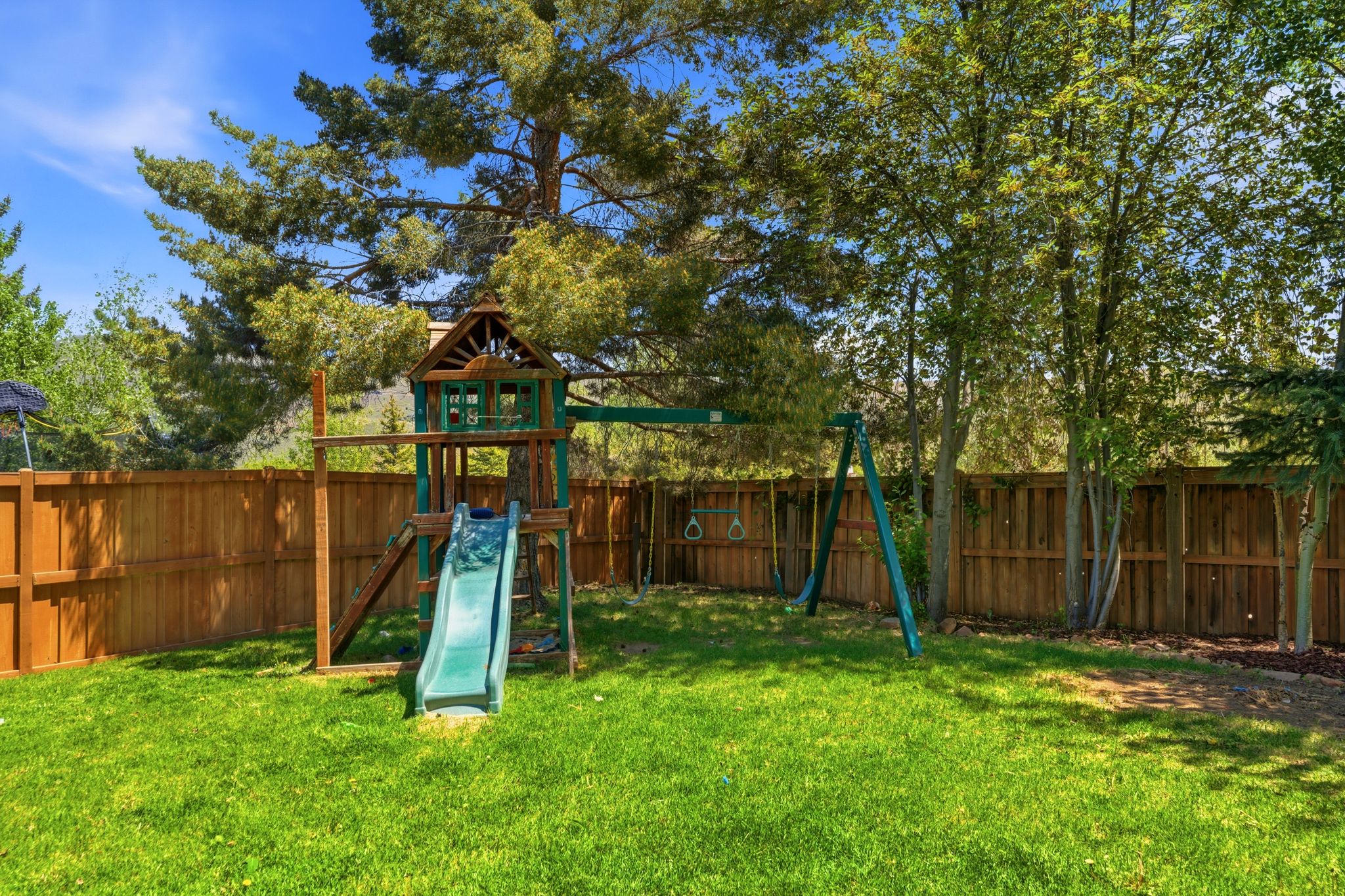 Expansive backyard featuring a lush green lawn, full privacy fencing, mature trees, and a dedicated play structure with slide and swings