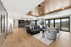 Spacious open-concept living area featuring light wood-finish flooring, a wood-paneled ceiling with recessed lighting, and expansive glass sliding doors
