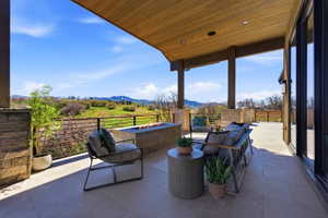 Expansive outdoor patio featuring a wood-finish ceiling, stone columns, large format tile flooring, and a linear built-in fire pit
