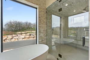 Freestanding soaking tub with a large picture window, a glass-enclosed shower with stone tile walls, a rain showerhead, and a rectangular window