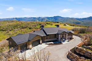 Modern mountain architecture featuring a dark asphalt shingle roof, stone and wood panel siding, an attached multi-car garage, and a paved driveway with a stone retaining wall