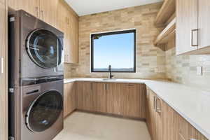 Dedicated laundry area featuring a stacked appliance pair, wood-finish cabinetry, light-toned countertops, and a rectangular window above a sink with a black faucet