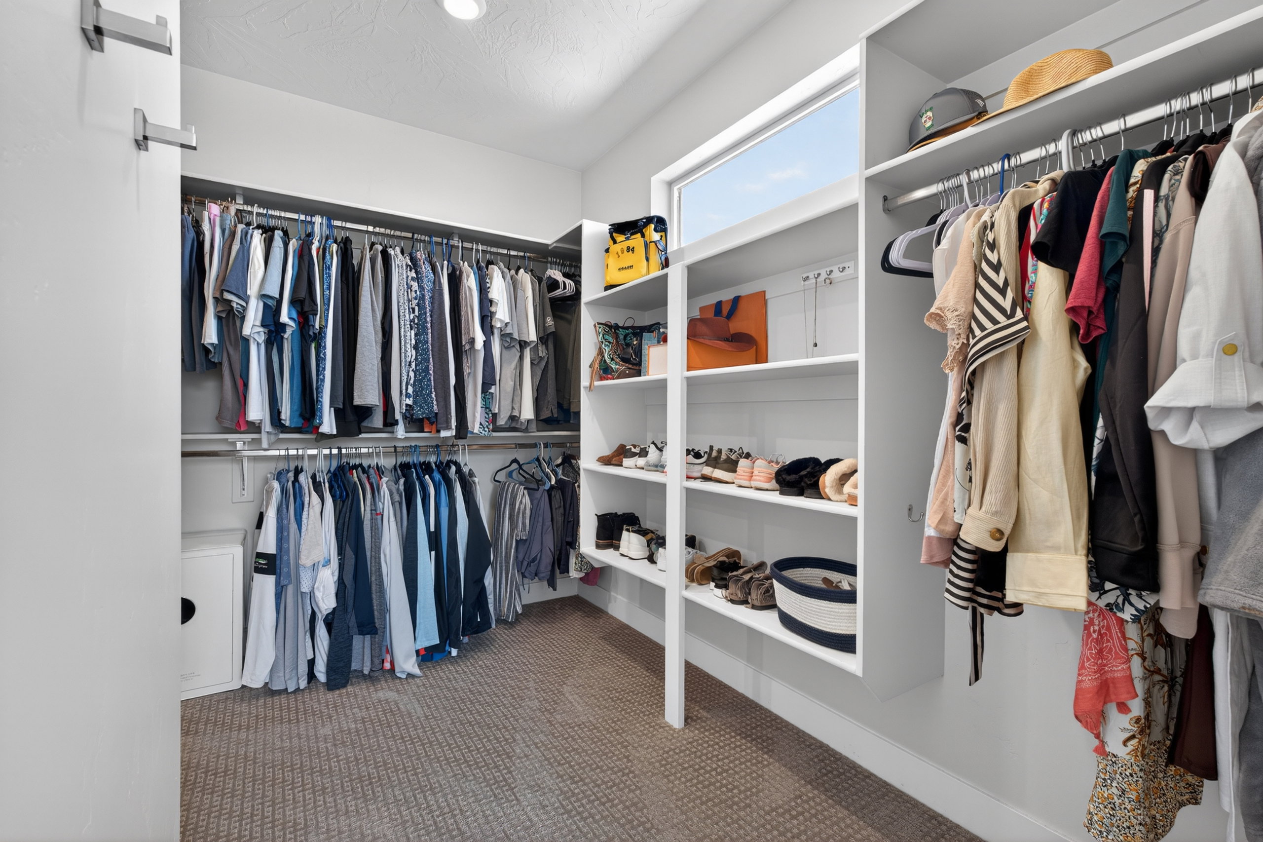 Spacious walk-in closet featuring white built-in shelving, chrome hanging rods, a high horizontal window, and patterned carpeting