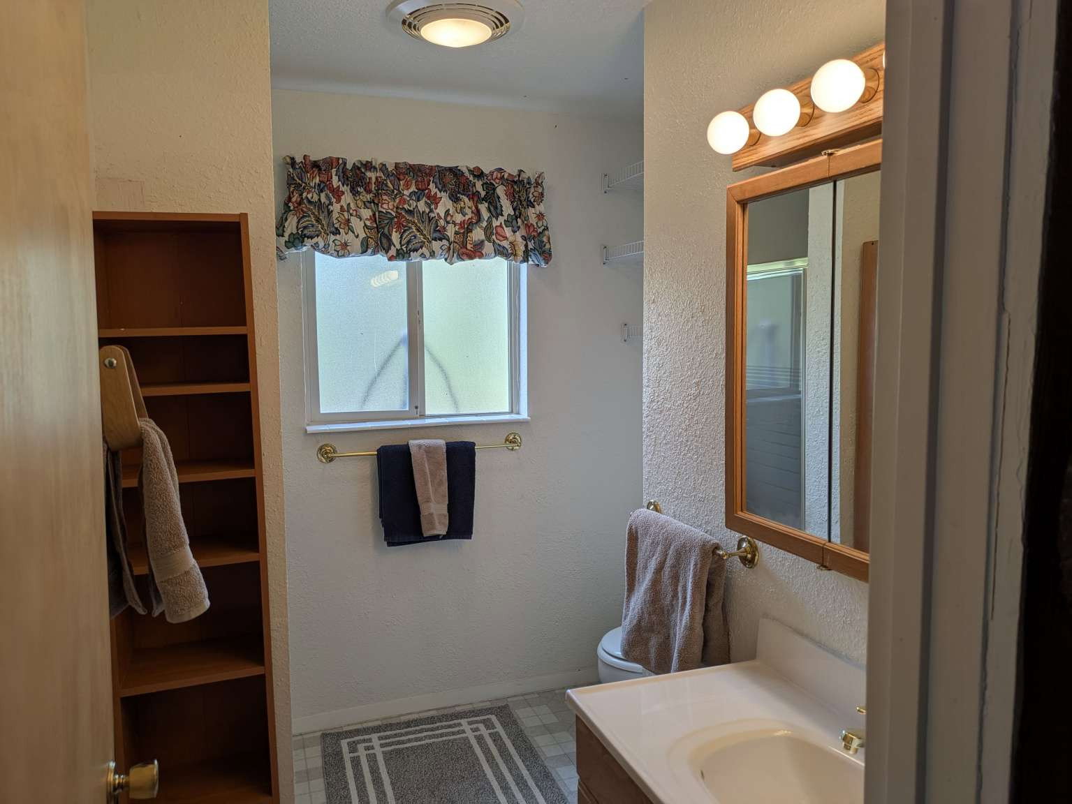 Built-in wood-finish shelving, single vanity with integrated sink, brass-finish faucet, framed mirror with overhead vanity lighting, and a frosted window