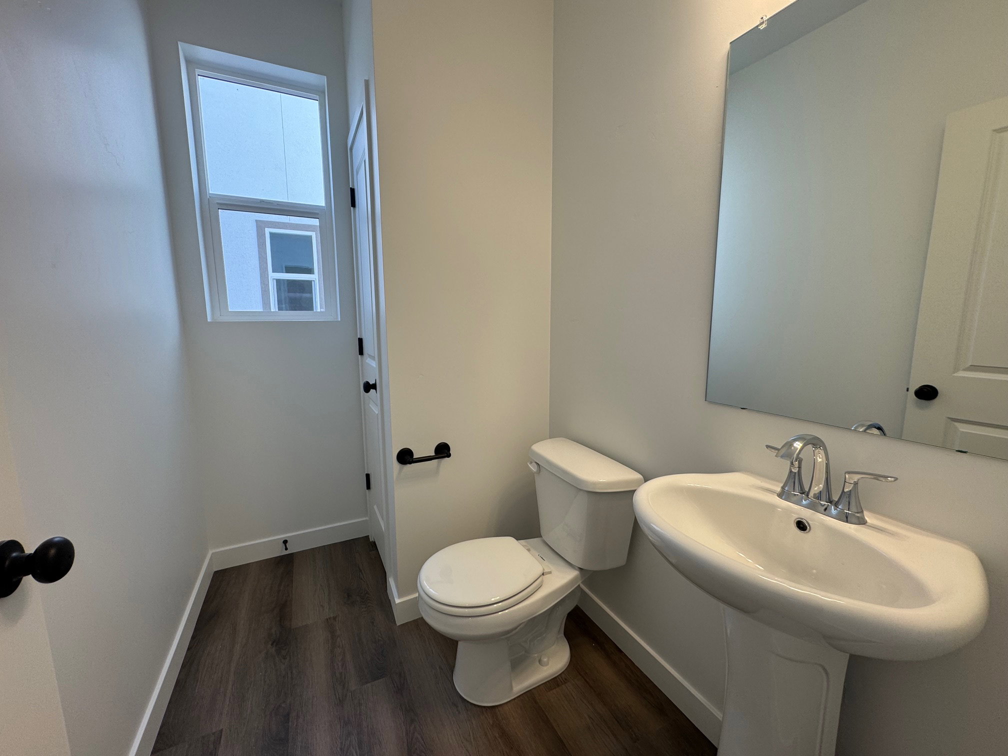 Bathroom featuring wood-finish flooring, a white pedestal sink with chrome fixtures, a rectangular wall-mounted mirror, a two-piece toilet, and a window for natural light