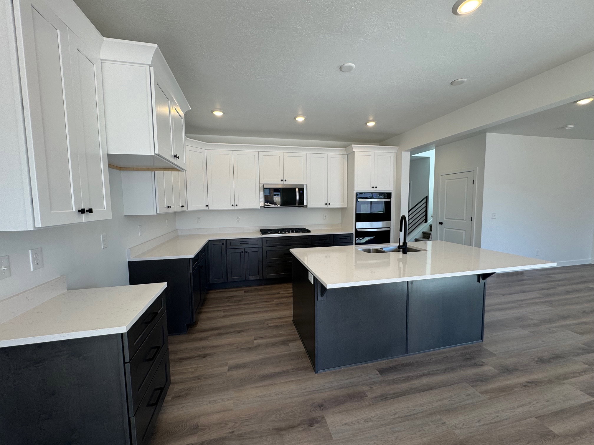 Spacious kitchen featuring two-tone cabinetry with white upper cabinets and dark lower cabinets, a central island with a white countertop and integrated sink, stainless steel appliances, and wood-finish flooring