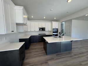 Spacious kitchen featuring two-tone cabinetry with white upper cabinets and dark lower cabinets, a central island with a white countertop and integrated sink, stainless steel appliances, and wood-finish flooring