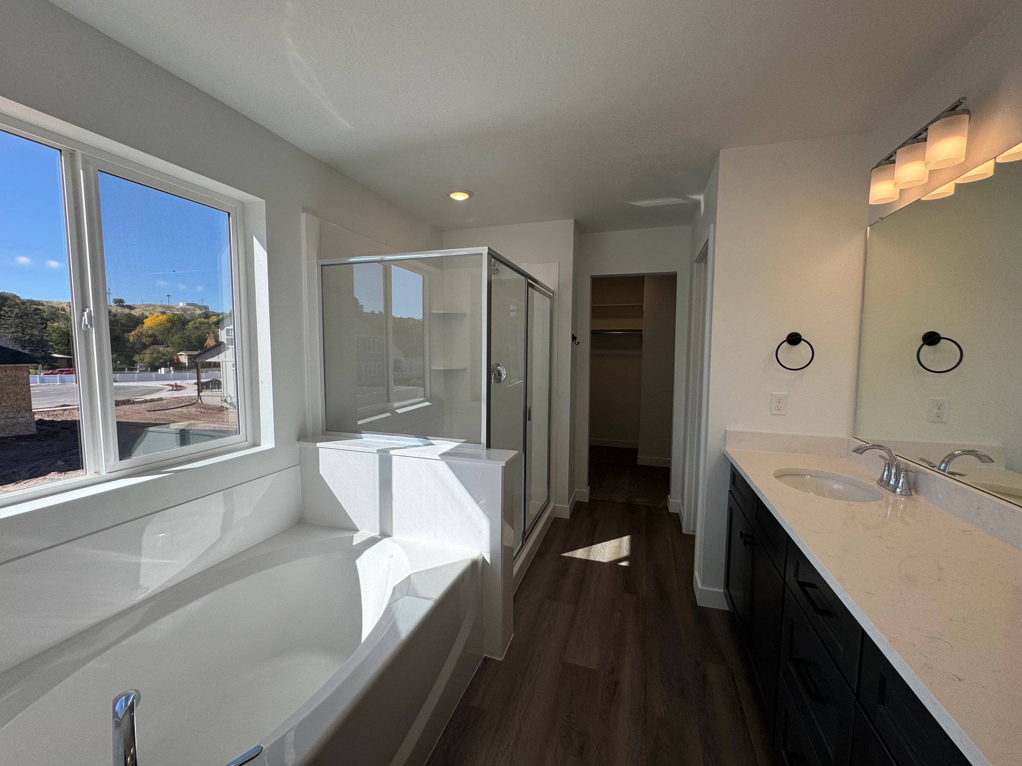 Bathroom featuring a built-in tub with surrounding paneling, a glass-enclosed shower, dual vanity with a light-toned countertop, a large mirror, and dark wood-finish flooring