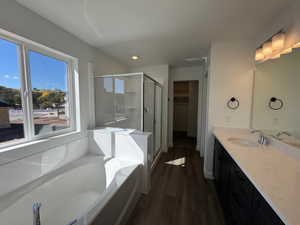 Bathroom featuring a built-in tub with surrounding paneling, a glass-enclosed shower, dual vanity with a light-toned countertop, a large mirror, and dark wood-finish flooring