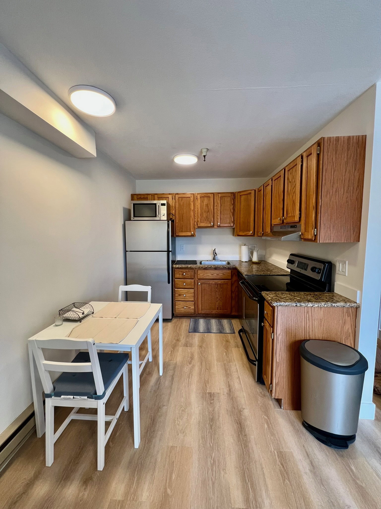 Compact kitchen featuring wood-finish cabinetry, granite-style countertops, stainless steel refrigerator, and a full-size electric range
