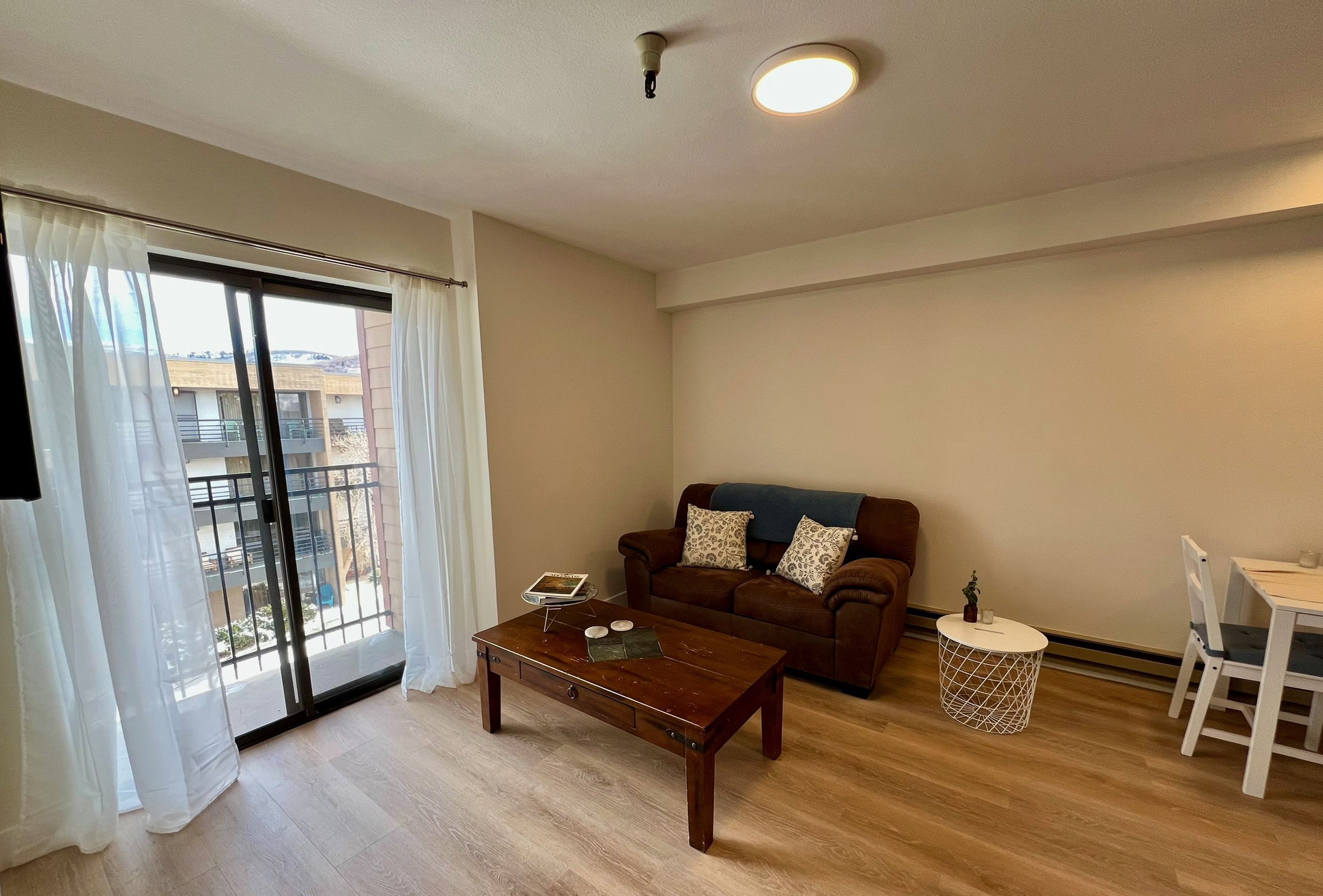 Spacious living area featuring light wood-finish flooring, neutral wall tones, and a sliding glass door leading to a private balcony with black metal railings